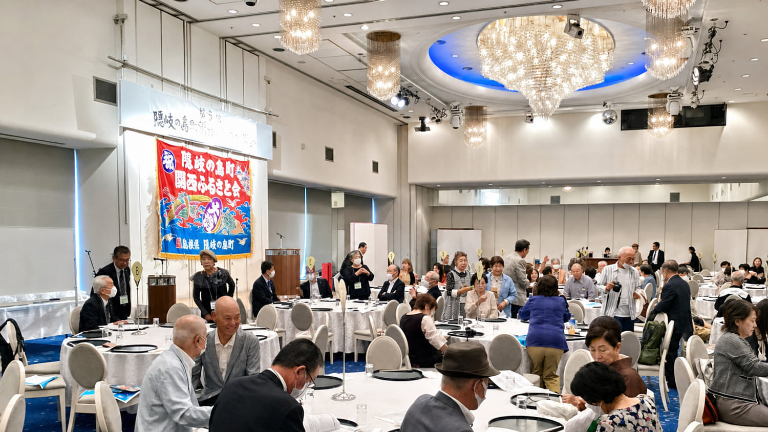 隠岐の島町関西ふるさと会 総会のようす。会旗が掲げられた会場に集う会員たち